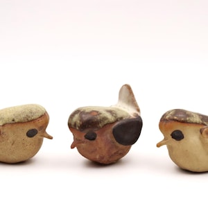 Könnte beinhalten: Drei Keramik-Vogelfiguren mit brauner, weißer und schwarzer Glasur. Die Vögel stehen in einer Reihe und blicken nach vorne.