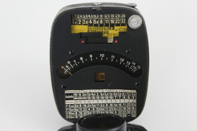 Puede incluir: Un fot&oacute;metro negro y plateado con un dial circular y una etiqueta amarilla con el texto "1.7 2.4 5.5 4 5.6 8 11 16 22 32 128 64 32 16 8 - CIN 96 16 10 10 10". El dial tiene marcas para los diafragmas y las velocidades de obturaci&oacute;n. El fot&oacute;metro tambi&eacute;n tiene una peque&ntilde;a ventana para ver la luz.