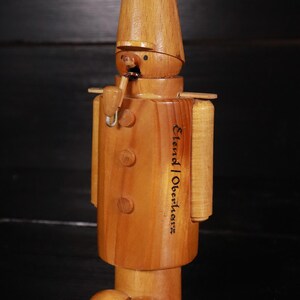 Könnte beinhalten: Holzfigur eines Mannes mit Hut und Pfeife. Die Figur ist aus hellem braunem Holz gefertigt und hat ein bemaltes Gesicht. Der Text "Elend Oberholz" ist auf den Körper der Figur eingraviert.