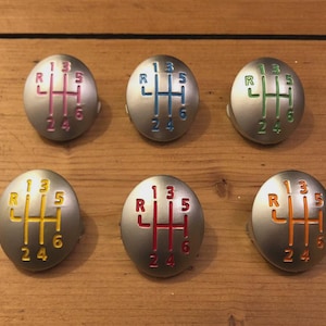 Könnte beinhalten: Sechs silberfarbene Schalthebelknöpfe mit farbigen Schaltmustern. Jeder Knopf hat ein anderes Farbschema: Pink, Blau, Grün, Gelb, Rot und Orange. Die Schaltmuster umfassen die Zahlen 1, 2, 3, 4, 5 und 6 sowie den Buchstaben R.