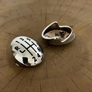 Könnte beinhalten: Zwei silberfarbene Schalthebelknöpfe für Autos. Ein Knopf ist oval mit einem schwarzen Schaltschema und Zahlen. Der andere Knopf ist eine silberfarbene, offen gehaltene Kappe. Beide liegen auf einer braunen Holzmaserung.