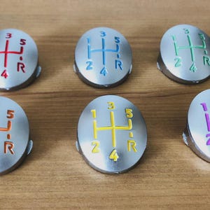 Puede incluir: Seis pomos de palanca de cambios plateados con patrones de cambio de color. Cada pomo tiene un color diferente para el patrón de cambio: rojo, azul, verde, naranja, amarillo y morado. Los pomos tienen forma ovalada.
