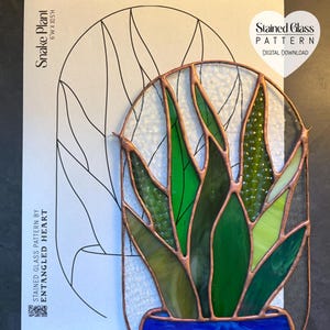 Pode incluir: Uma planta de serpente em vitral num vaso azul com linhas de soldadura de cobre, exibida sobre um padrão de papel branco com a etiqueta Snake Plant 6"W x 10.5"H e Stained Glass Pattern by Entangled Heart com um ícone de coração.