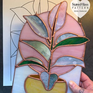 Op de afbeelding: Een Ficus plant zonnevanger van glas-in-lood met roze, groene en gele glazen onderdelen verbonden door koperfolie soldeerlijnen. Het stuk wordt getoond voor een papieren patroon met het label Ficus Plant 7,25 inch B x 10,5 inch H.