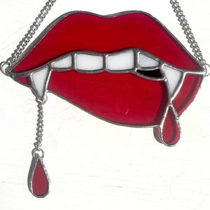 May include: A stained glass piece depicting a pair of red lips with white teeth and two fangs. A silver chain is attached to the top for hanging, with a red teardrop hanging from the bottom.