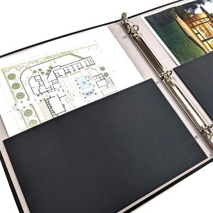May include: A black three-ring binder with a silver metal ring mechanism. The binder has two black pockets, one on each side, and is open to reveal a blueprint and a photograph.