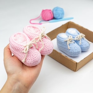 May include: Two pairs of crocheted baby booties, one pink and one blue, with white laces. The pink booties are being held in a hand, while the blue booties are in a cardboard box.