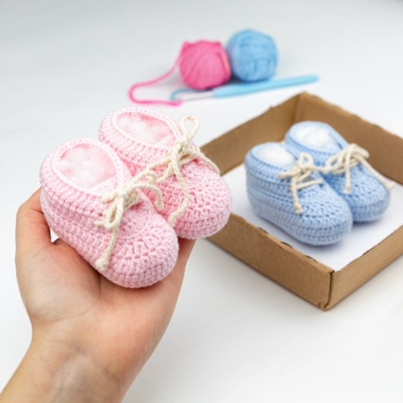 May include: Two pairs of crocheted baby booties, one pink and one blue, with white laces. The pink booties are being held in a hand, while the blue booties are in a cardboard box.