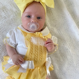 May include: A baby wearing a yellow knitted romper with a white bow and a yellow bow headband. The romper has a white lace trim and a white collar. The baby is wearing a pacifier.