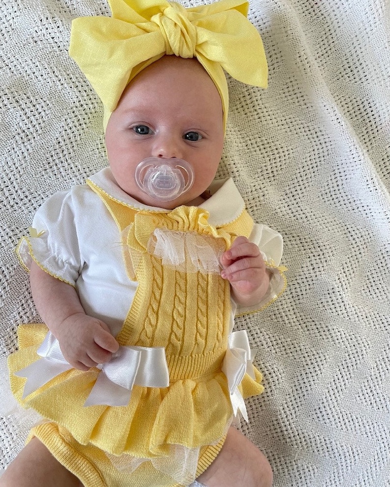 May include: A baby wearing a yellow knitted romper with a white bow and a yellow bow headband. The romper has a white lace trim and a white collar. The baby is wearing a pacifier.