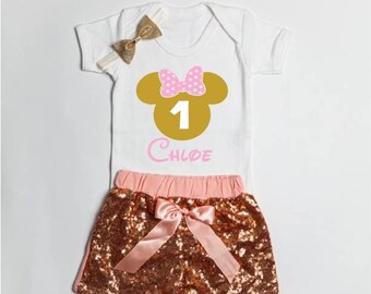 May include: A white short-sleeved bodysuit featuring a gold mouse-ear silhouette with a pink polka-dot bow, the number 1, and the name Chloe in pink script. Includes matching gold sequined shorts with a pink satin bow and a gold hair bow.