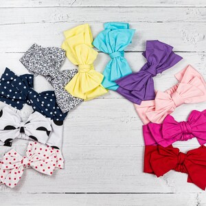 May include: Assortment of colorful fabric headbands with bow designs. The headbands are arranged in a semi-circle on a white wooden surface. The headbands are in various colors, including pink, red, blue, yellow, and purple.