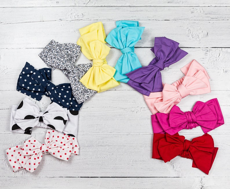 May include: Assortment of colorful fabric headbands with bow designs. The headbands are arranged in a semi-circle on a white wooden surface. The headbands are in various colors, including pink, red, blue, yellow, and purple.