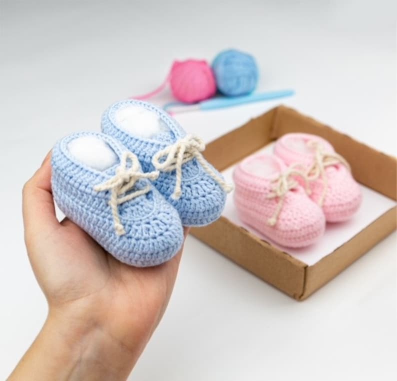 May include: Two pairs of crocheted baby shoes, one pair is blue and the other pair is pink. The shoes have white laces and are in a cardboard box.