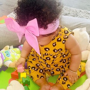 May include: A baby wearing a yellow and black leopard print jumpsuit with a pink bow headband. The baby is sitting on a green and orange play mat with colorful wooden blocks and stuffed animals.