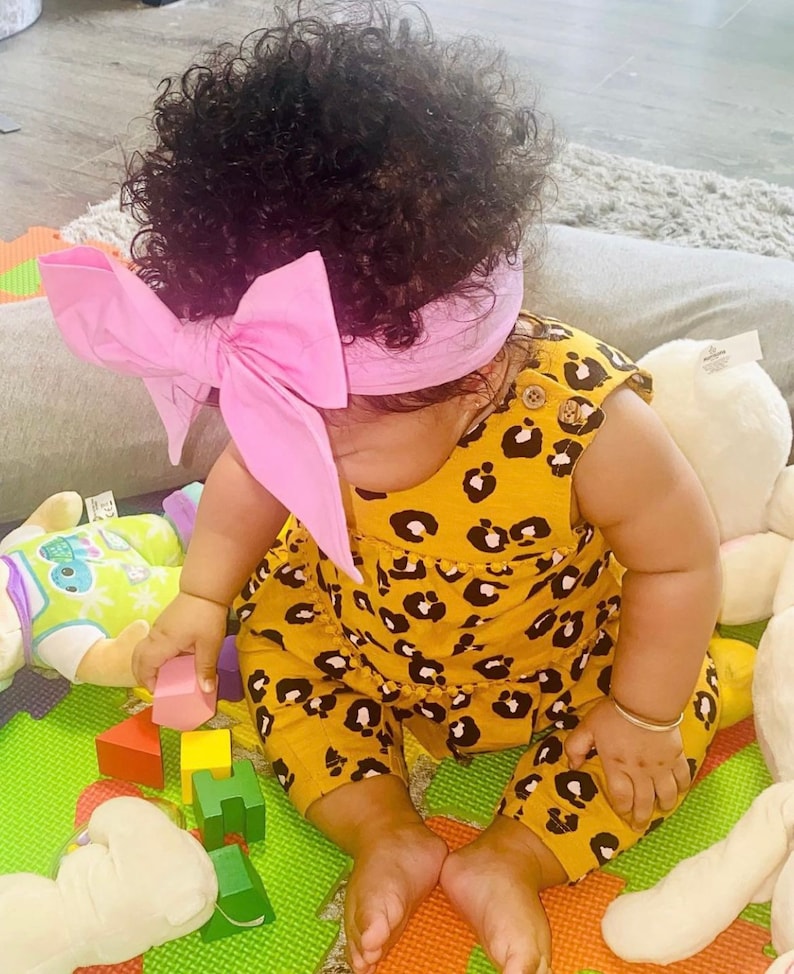 May include: A baby wearing a yellow and black leopard print jumpsuit with a pink bow headband. The baby is sitting on a green and orange play mat with colorful wooden blocks and stuffed animals.