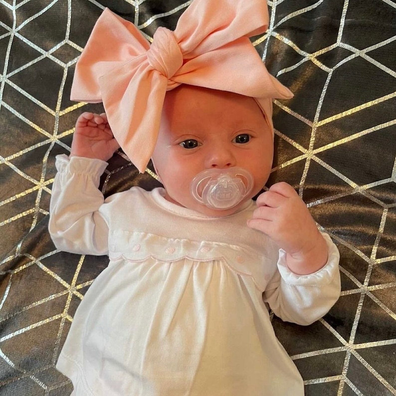 May include: A baby wearing a white long-sleeved dress with a pink bow headband and a pacifier in their mouth.