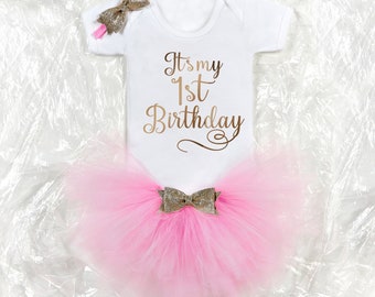 May include: A pink tulle tutu with a gold glitter bow, a white onesie with the text "It's my 1st Birthday" in gold glitter, and a gold glitter headband with a bow.