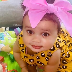 May include: A baby girl wearing a yellow and black leopard print jumpsuit with a pink bow headband. The baby is sitting on a green and yellow play mat.