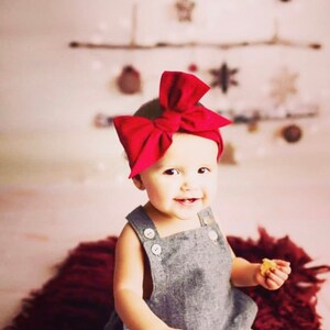May include: A baby girl wearing a gray overall dress with buttons and a large red bow headband. She is smiling and holding a small snack.