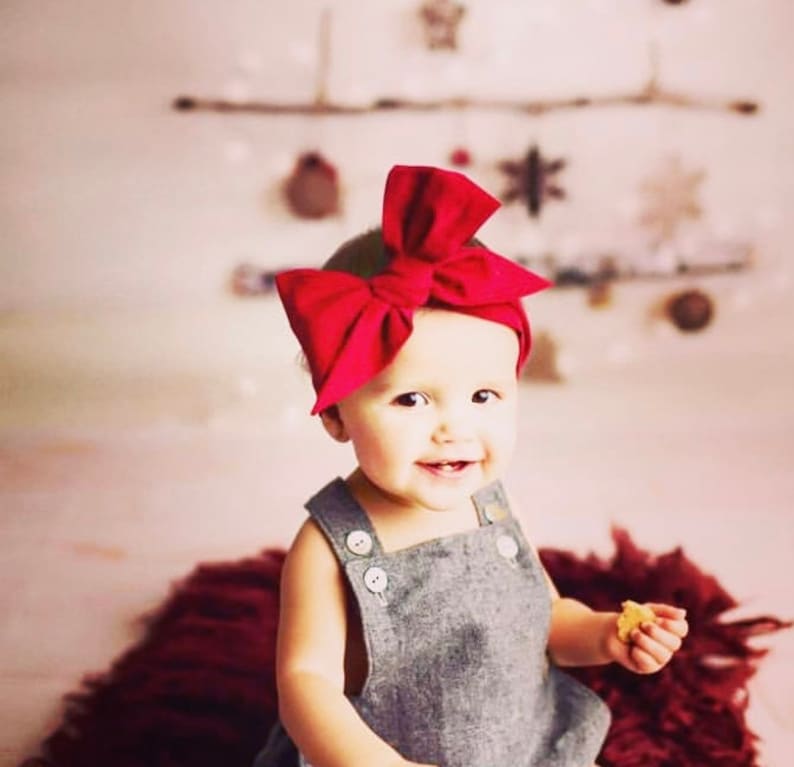 May include: A baby girl wearing a gray overall dress with buttons and a large red bow headband. She is smiling and holding a small snack.