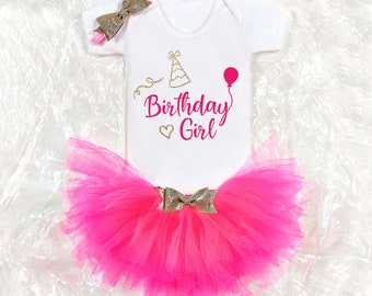 May include: A pink tulle tutu skirt with a gold bow and a white onesie with the text "Birthday Girl" in gold glitter and a pink party hat and balloon graphic.
