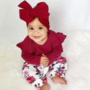 May include: A baby wearing a burgundy long-sleeved shirt with ruffled sleeves and white pants with a floral print. The baby is wearing a burgundy bow headband.