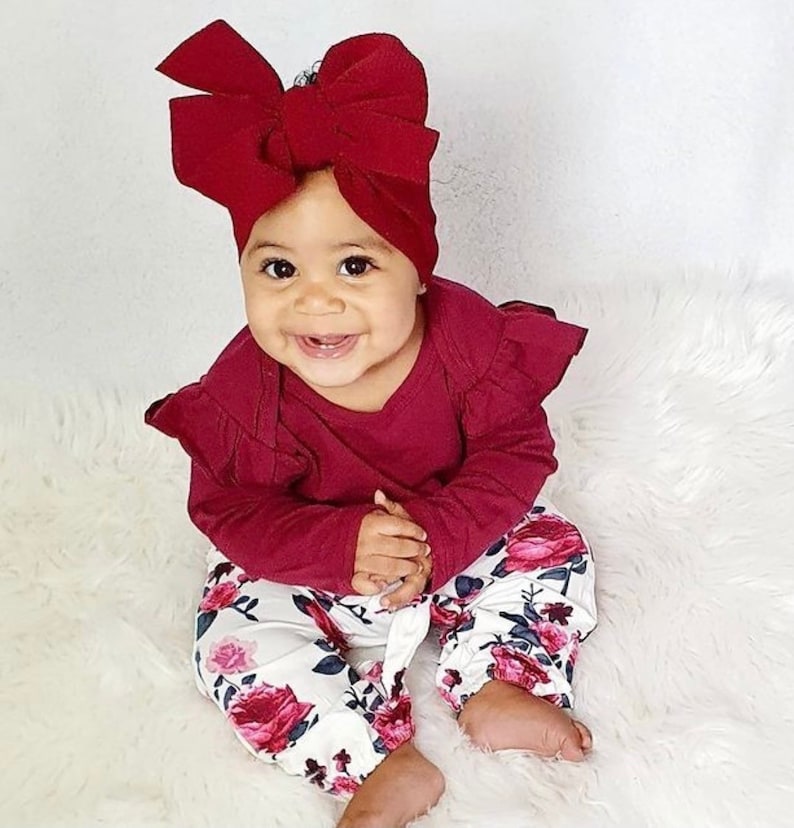 May include: A baby wearing a burgundy long-sleeved shirt with ruffled sleeves and white pants with a floral print. The baby is wearing a burgundy bow headband.