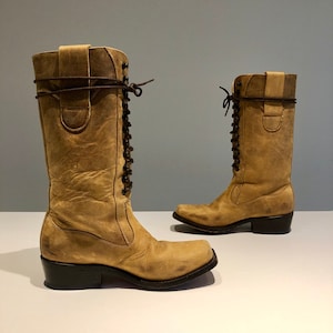 Puede incluir: Un par de botas de cuero color beige con cordones en la parte delantera y punta cuadrada. Las botas tienen un tacón bajo y están hechas de cuero desgastado.