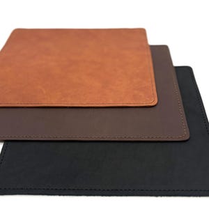 May include: Three leather desk pads in shades of brown and black are stacked on a white surface. The top pad is a light brown, the middle is a darker brown, and the bottom is black. Each pad has stitched edges.