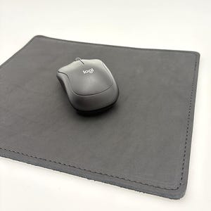 May include: A black computer mouse with the "logi" logo rests on a dark gray, rectangular leather mousepad. The mousepad has stitched edges and a smooth surface. The background is plain white.