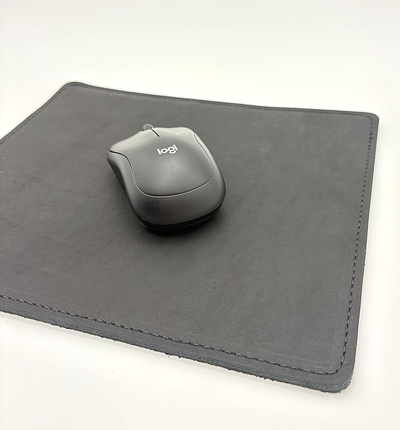 May include: A black computer mouse with the "logi" logo rests on a dark gray, rectangular leather mousepad. The mousepad has stitched edges and a smooth surface. The background is plain white.