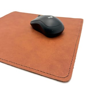 May include: A brown leather mousepad with a black wireless mouse. The mousepad is rectangular with stitched edges. The mouse has the Logitech logo.