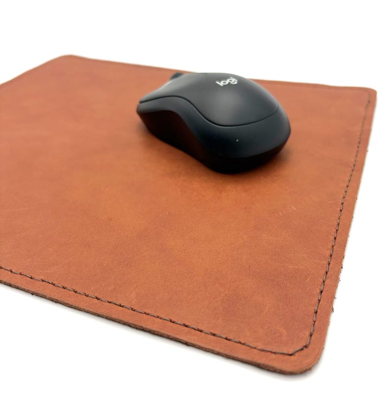 May include: A brown leather mousepad with a black wireless mouse. The mousepad is rectangular with stitched edges. The mouse has the Logitech logo.