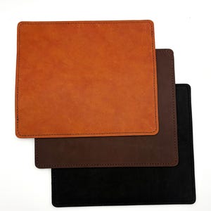 May include: Three square leather desk pads in shades of orange, brown, and black are stacked on a white background. Each pad has a stitched border. The top pad is a light orange color, the middle pad is brown, and the bottom pad is black.