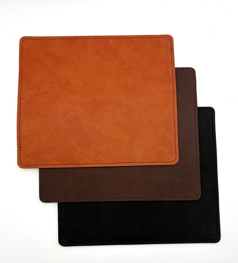 May include: Three square leather desk pads in shades of orange, brown, and black are stacked on a white background. Each pad has a stitched border. The top pad is a light orange color, the middle pad is brown, and the bottom pad is black.