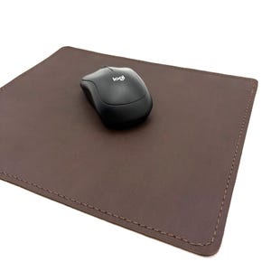 May include: A brown leather mouse pad with a black wireless mouse. The mouse pad is rectangular with stitched edges. The mouse has the "logi" logo.