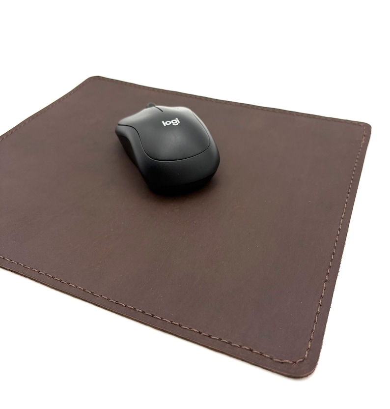 May include: A brown leather mouse pad with a black wireless mouse. The mouse pad is rectangular with stitched edges. The mouse has the "logi" logo.