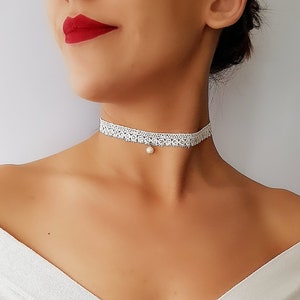 Może przedstawiać: Biały choker z koronki z pojedynczym wisiorem z pereł. Choker jest delikatny i kobiecy, a perła dodaje mu elegancji.