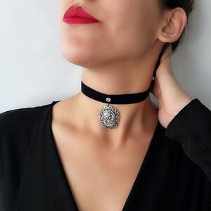 Op de afbeelding: Een zwarte fluwelen chokerketting met een zilveren hanger met een gedetailleerd ontwerp. De hanger heeft een kleine, heldere strasssteen in het midden.