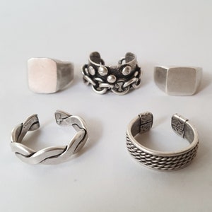 Puede incluir: Cuatro anillos de plata, dos con un diseño cuadrado simple, uno con un diseño trenzado y uno con un diseño de eslabón de cadena.