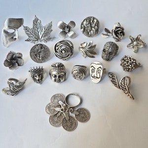 May include: A collection of silver-toned rings featuring various designs, including a flower, a skull, a cat, a face, and a feather. The rings are arranged on a white surface.