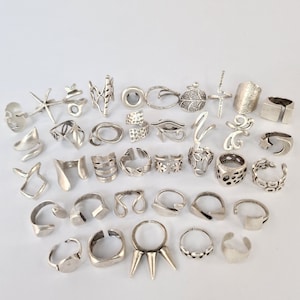 May include: A collection of 30 silver rings with various designs, including geometric shapes, floral patterns, and abstract forms. The rings are arranged in a semi-circle on a white background.