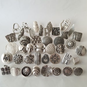 May include: A collection of silver-toned rings with various designs, including geometric patterns, floral motifs, and abstract shapes. The rings are arranged on a white surface.