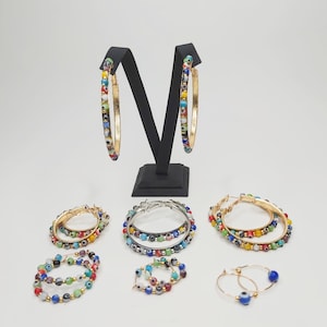 Könnte beinhalten: Ein Set mit sechs goldenen Creolen mit bunten bösen Augenperlen. Die Ohrringe sind auf einem schwarzen Ständer und einer weißen Oberfläche angeordnet.