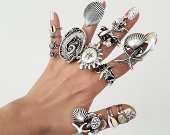 Könnte beinhalten: Ein Handmodell trägt eine Vielzahl von silbernen Ringen mit Meeresmotiven, darunter Muscheln, Seesterne und eine Sonne. Die Ringe sind auf mehreren Fingern übereinander getragen.
