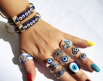 May include: A hand wearing two leather bracelets with blue evil eye charms and multiple silver rings with blue evil eye stones. The rings are on different fingers and some have a white background.