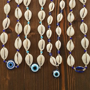 Puede incluir: Cinco collares de conchas blancas con cuentas azules y turquesas y un amuleto azul de ojo turco. Los collares están sobre una superficie de madera marrón.