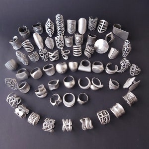 May include: A collection of silver-toned rings with various designs, including geometric, floral, and abstract patterns. The rings are arranged in a circular pattern on a dark gray background.