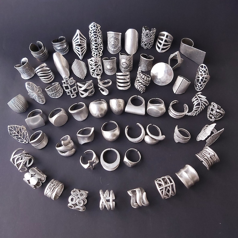 May include: A collection of silver-toned rings with various designs, including geometric, floral, and abstract patterns. The rings are arranged in a circular pattern on a dark gray background.